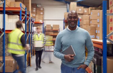 Portrait Of Manager With Digital Tablet In Busy Modern Warehouse With Staff In Background VMI to mitigate geopolitical risk in supply chain